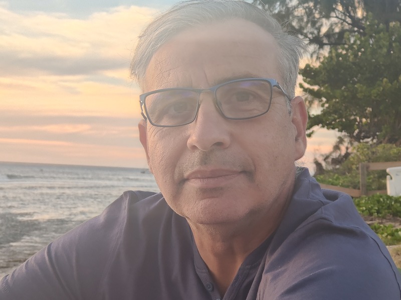 photo de profil de Bertrand Tanguy. Prise en bordure de mer avec en arrière plan a droite une zone arboré et a gauche de Bertrand la mer. Bertrand porte des lunettes et un polo bleu. la photo à été prise le soir lors du couché de soleil.